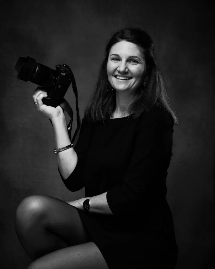 Portrait corporate d’une professionnelle de la photographie, sourire naturel en studio noir et blanc.