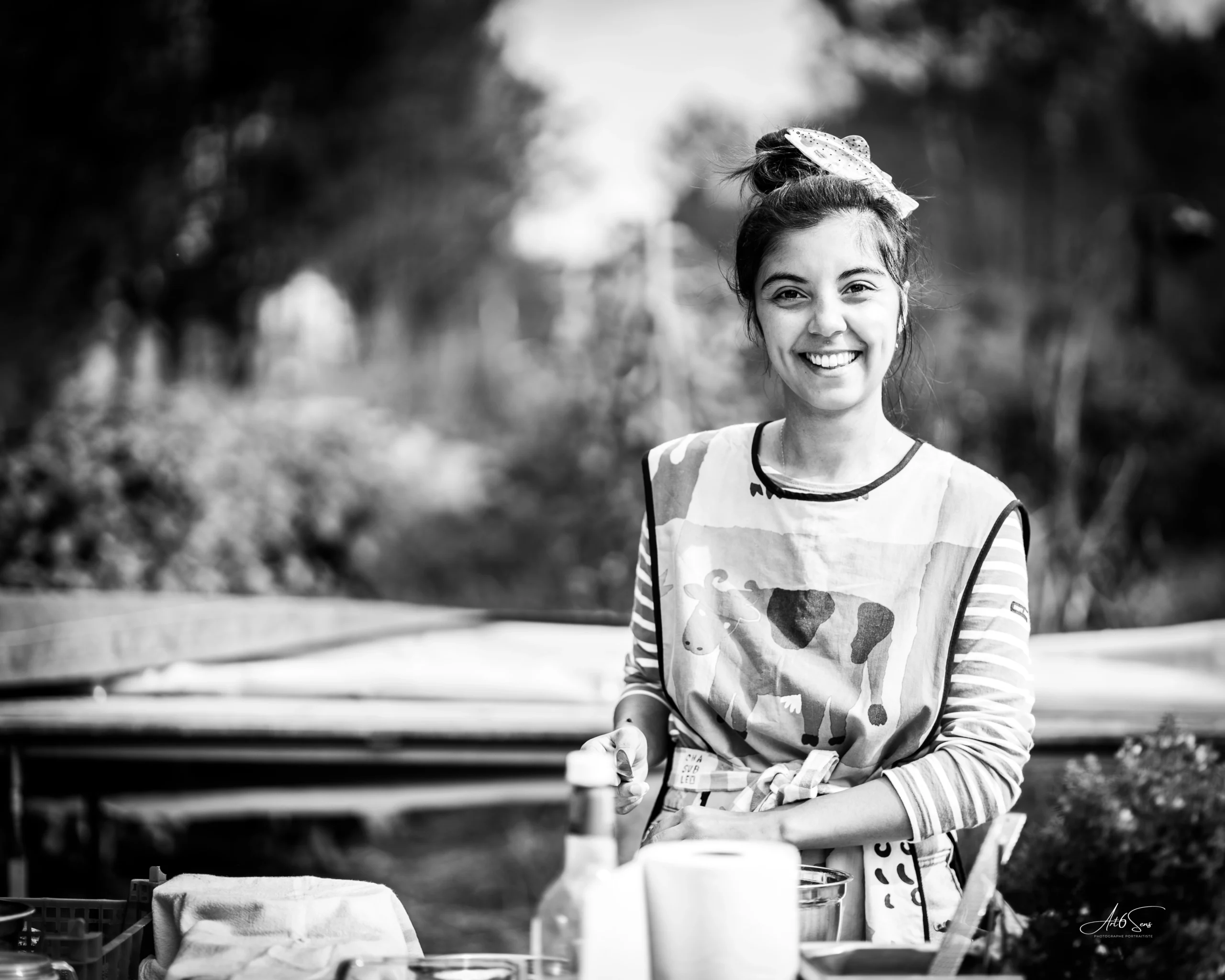 Portrait en noir et blanc d’une femme souriante portant un tablier, photographie naturelle réalisée en extérieur à La Teste par Art6Sens, Portraitiste de France.